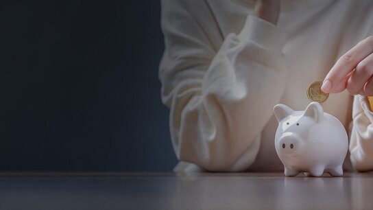 Hände stecken eine Münze in ein weißes Sparschwein auf einem Tisch in einem schummrigen Raum.