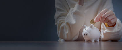 Hände stecken eine Münze in ein weißes Sparschwein auf einem Tisch in einem schummrigen Raum.