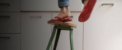 Eine Person steht auf einem Holzschemel, um etwas in einer Küchenzeile zu erreichen, trägt rote Sandalen und Jeans.
