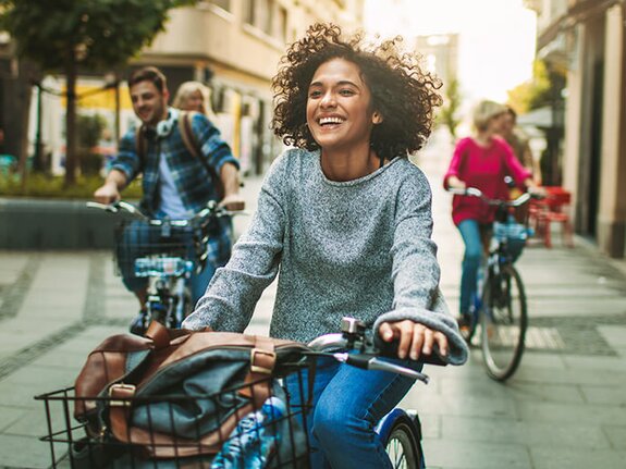 Fahradfahrer in gelber Jacke fährt über einen Kopfsteinpflasterweg bei Sonnenuntergang, im Hintergrund moderne Gebäude.