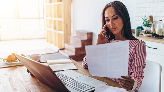 Junge Frau sitzt am Tisch, telefoniert und liest ein Dokument, während ein Laptop und eine Tasse daneben stehen.