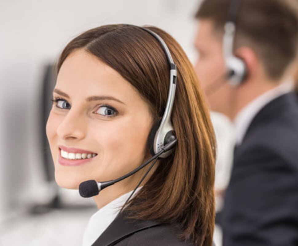 Frau mit Kopfhörern und Mikrofon lächelt zufrieden in einem Callcenter, im Hintergrund weitere Mitarbeiter.