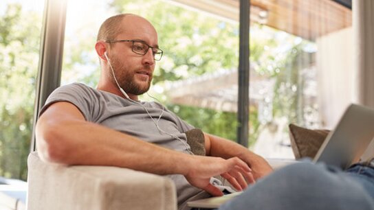 Mann mit Kopfhörern sitzt entspannt auf einem Sofa und arbeitet konzentriert am Laptop. Helles, freundliches Ambiente.
