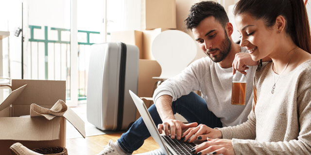 Junge Frau und Mann sitzen auf dem Boden, sie arbeitet am Laptop, er hält ein Getränk, umgeben von Umzugskartons.