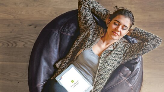 Frau sitzt entspannt in einem Ohrensessel, hält ein Tablet und schließt die Augen. Helle Holzoberfläche im Hintergrund.