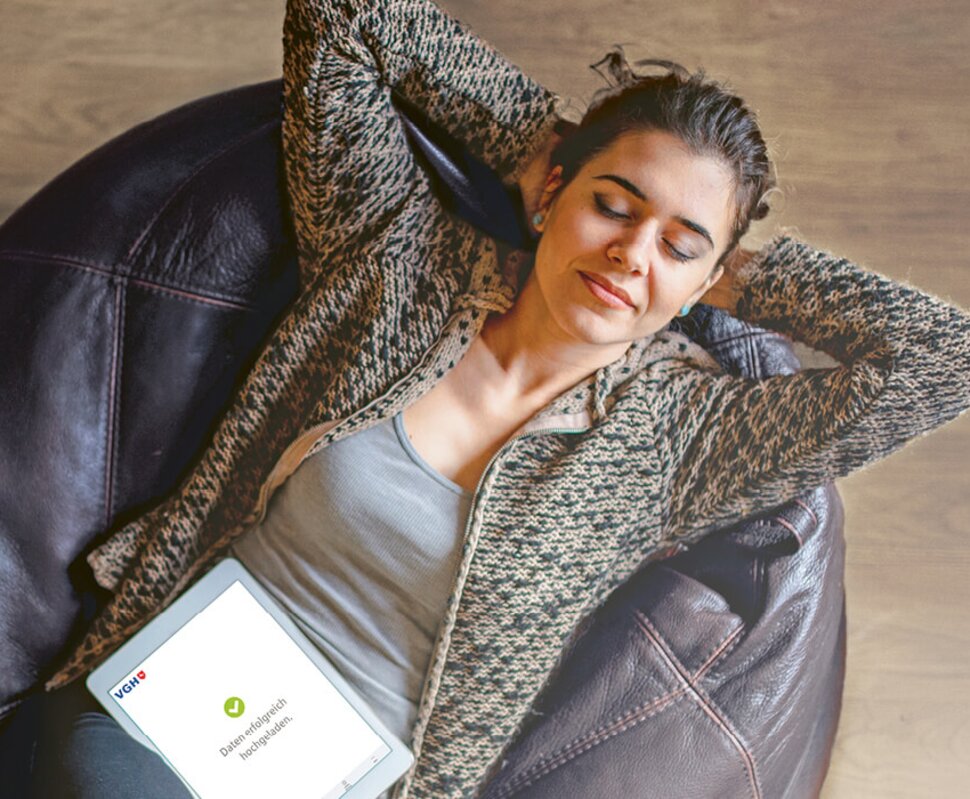 Frau sitzt entspannt in einem Ohrensessel, hält ein Tablet und schließt die Augen. Helle Holzoberfläche im Hintergrund.