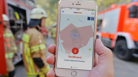 Smartphone zeigt eine Kartenansicht mit der Markierung "Großbrand" im Einsatzfeld, Feuerwehrleute im Hintergrund.