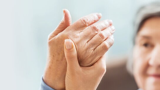 Zwei Hände halten sich sanft, eine ältere Hand wird von einer jüngeren Hand unterstützt. Hintergrund unscharf.