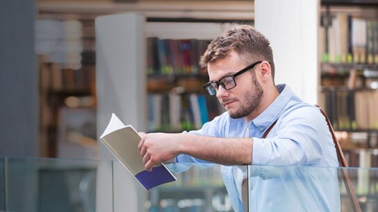 Junger Mann mit Brille liest konzentriert ein Buch in einer modernen Bibliothek. Bücherregale im Hintergrund sichtbar.