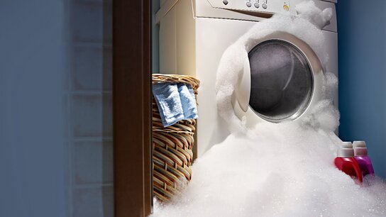 Waschmaschine, umgeben von Schaum, daneben ein Wäschekorb mit Kleidung und zwei Flaschen Waschmittel.