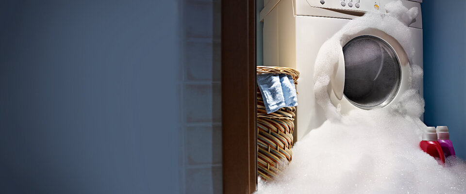 Waschmaschine, umgeben von Schaum, daneben ein Wäschekorb mit Kleidung und zwei Flaschen Waschmittel.