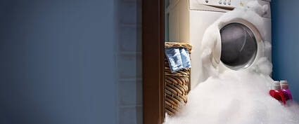 Waschmaschine, umgeben von Schaum, daneben ein Wäschekorb mit Kleidung und zwei Flaschen Waschmittel.