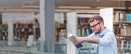 Junger Mann liest ein Buch in einer modernen Bibliothek, umgeben von Bücherregalen und hellem Licht.