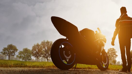 Mann mit Helm, der von einer Sportmotorrad auf einer ländlichen Straße ins Sonnenlicht geht, umgeben von Bäumen.