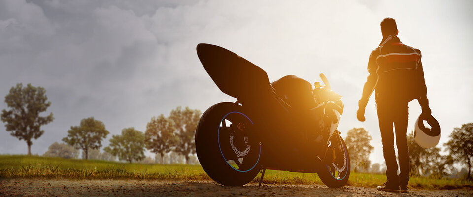 Mann mit Helm, der von einer Sportmotorrad auf einer ländlichen Straße ins Sonnenlicht geht, umgeben von Bäumen.