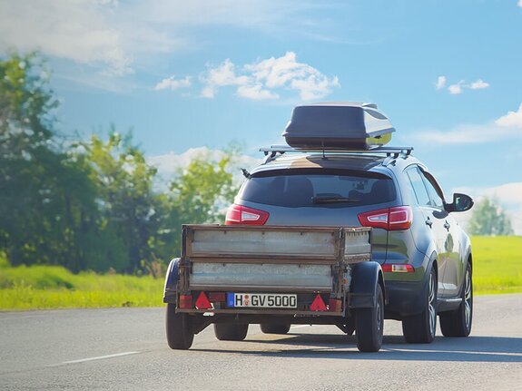 Ein SUV mit Anhänger auf einer Landstraße, umgeben von grünen Bäumen und klarem Himmel.