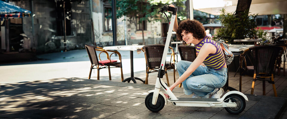 Eine Person mit lockigem Haar sitzt auf einem weißen Elektroroller in einem Straßencafé.