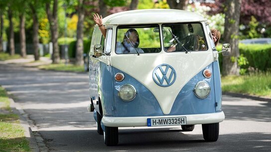 Ein Volkswagen-Bus fährt eine ruhige Straße entlang, Fahrer und Beifahrer winken fröhlich aus den Fenstern.