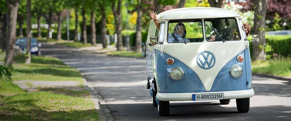 Ein Volkswagen-Bus fährt eine ruhige Straße entlang, Fahrer und Beifahrer winken fröhlich aus den Fenstern.