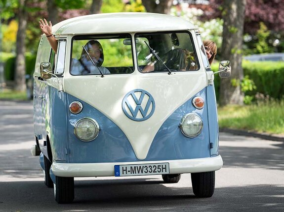 Ein Volkswagen-Bus fährt eine ruhige Straße entlang, Fahrer und Beifahrer winken fröhlich aus den Fenstern.