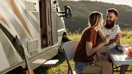 Paar sitzt im Freien vor einem Wohnmobil, genießt Snacks und Getränke, umgeben von Natur. Entspannte Stimmung.