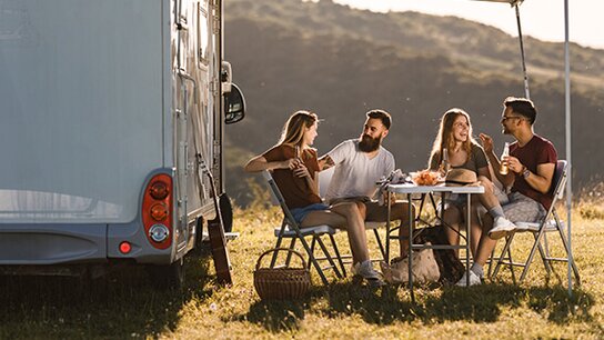 Vier Freunde sitzen entspannt an einem Tisch vor einem Wohnmobil und genießen Snacks in einer sonnigen, ländlichen Umgebung.