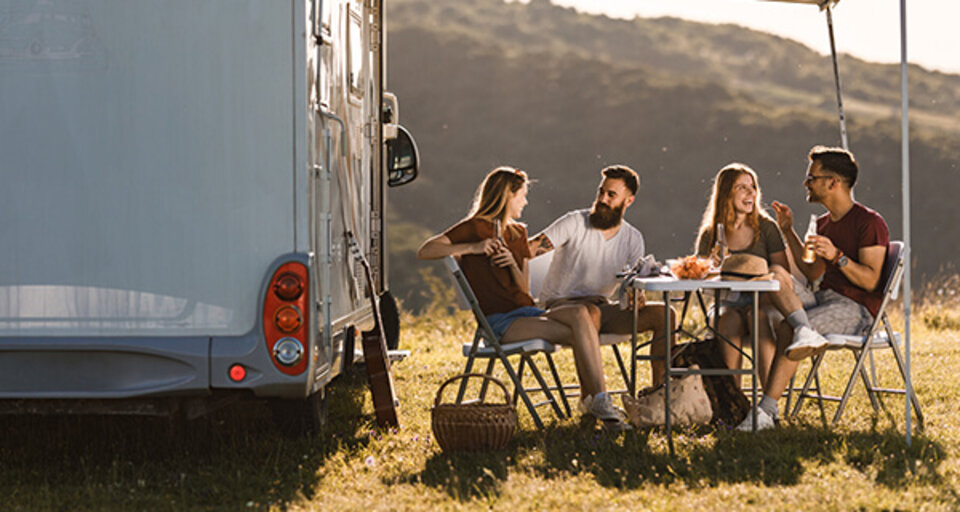 Vier Freunde sitzen entspannt an einem Tisch vor einem Wohnmobil und genießen Snacks in einer sonnigen, ländlichen Umgebung.