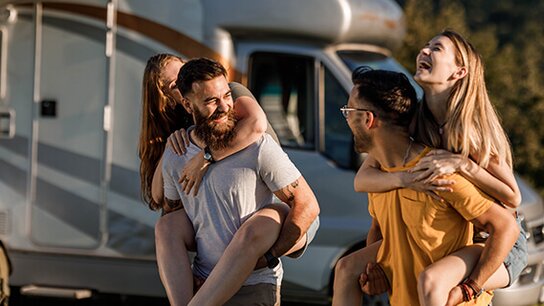 Vier Freunde lachen und haben Spaß, während zwei Frauen auf den Schultern ihrer männlichen Freunde vor einem Wohnmobil sitzen.