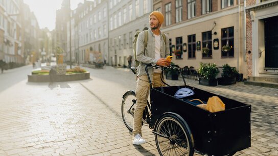 Mann mit Beanie und Rucksack steht neben einem Lastenrad in einer sonnigen, ruhigen Straßenansicht.