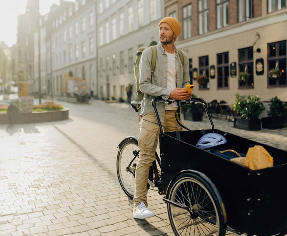 Mann mit Beanie und Rucksack steht neben einem Lastenrad in einer sonnigen, ruhigen Straßenansicht.