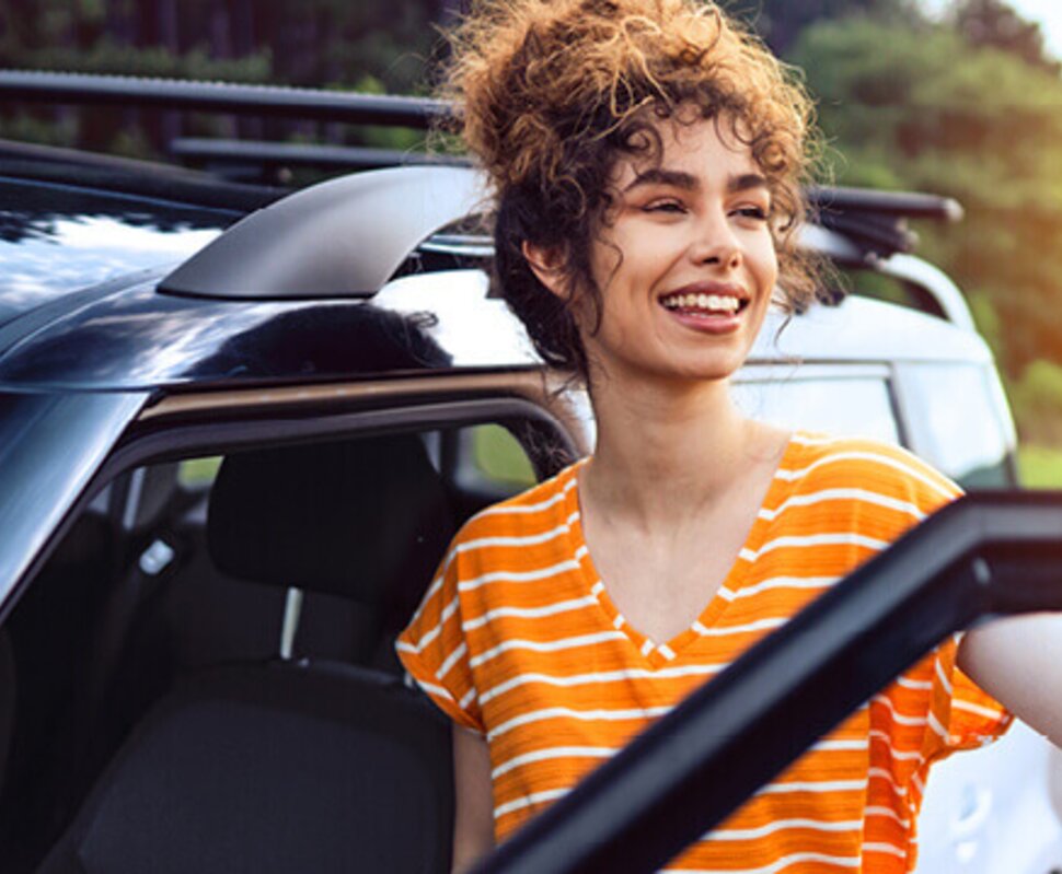Lächelnde Frau mit lockigem Haar, die aus einem Auto steigt, im Hintergrund Bäume und ein weiteres Fahrzeug.