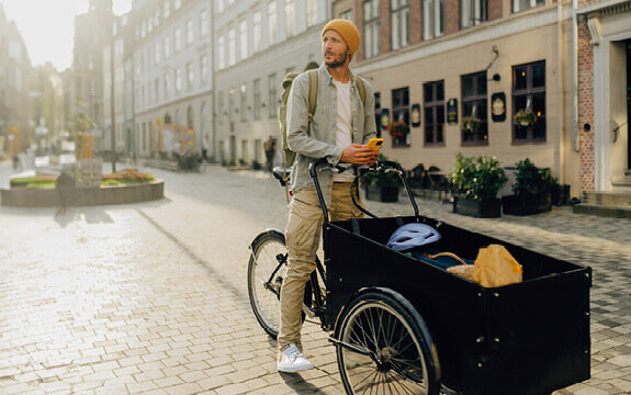 Mann mit orangefarbener Mütze steht an einem Lastenrad in einer ruhigen, sonnigen Straßenumgebung.