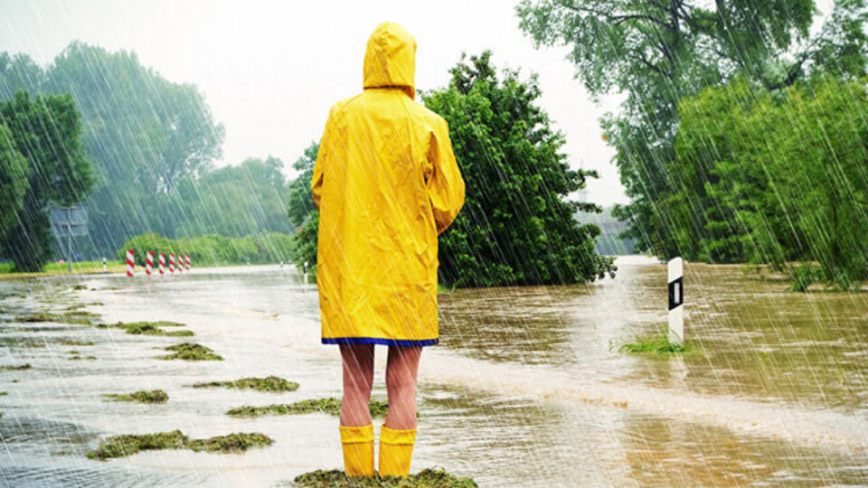 Eine Person in gelbem Regenmantel steht auf einer überschwemmten Straße, während starker Regen fällt.