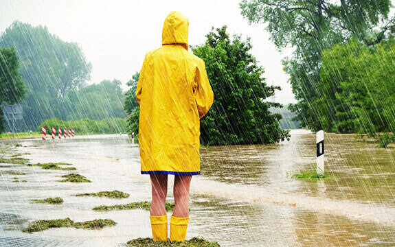 Eine Person in gelbem Regenmantel steht auf einer überschwemmten Straße, während starker Regen fällt.