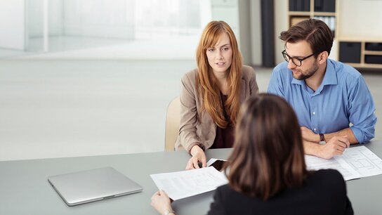 Drei Personen in einem modernen Büroraum sitzen an einem Tisch, während sie über Dokumente diskutieren.