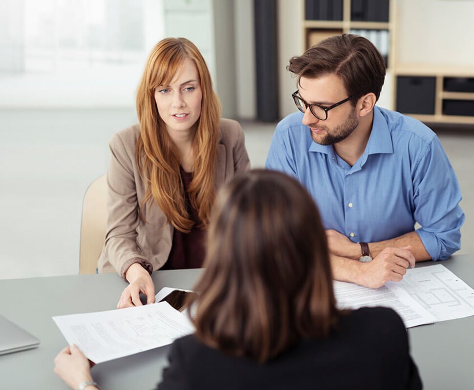 Drei Personen in einem modernen Büroraum sitzen an einem Tisch, während sie über Dokumente diskutieren.