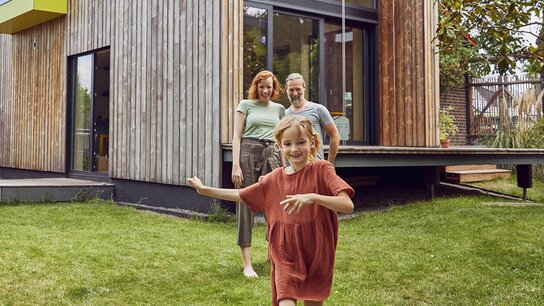 Eine Familie mit einem Mädchen, das lachend im Garten läuft, steht vor einem modernen Holzhaus.