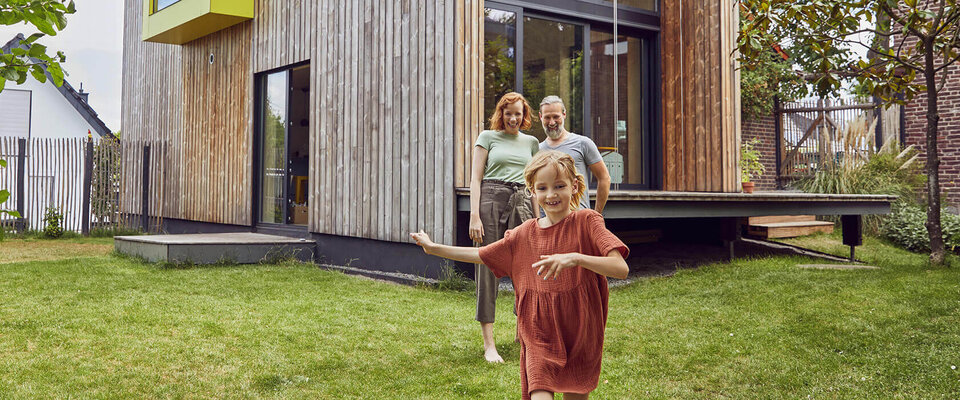 Eine Familie mit einem Mädchen, das lachend im Garten läuft, steht vor einem modernen Holzhaus.