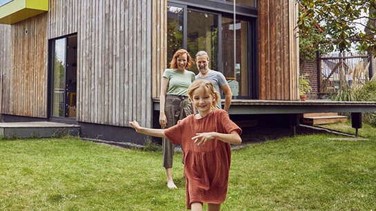 Familie im Garten vor einem modernen Holzhaus; ein Mädchen läuft fröhlich voraus, während die Eltern lächelnd beobachten.