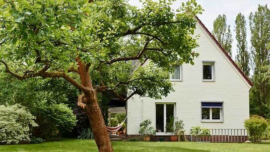 Weißes Einfamilienhaus mit Garten, umgeben von Bäumen und bepflanzten Beeten, sowie einer Hängematte im Hintergrund.