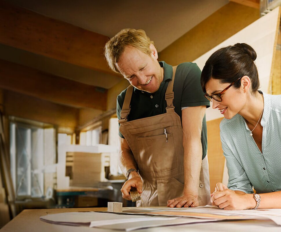 Ein Handwerker und eine Frau betrachten Pläne auf einem Tisch in einer Werkstatt, umgeben von Materialien und Werkzeugen.