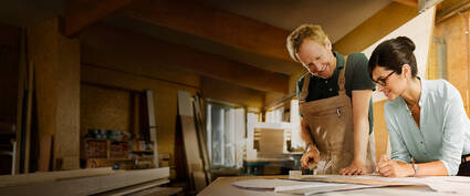 Ein Handwerker und eine Frau betrachten Pläne auf einem Tisch in einer Werkstatt, umgeben von Materialien und Werkzeugen.
