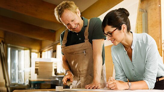 Ein Handwerker und eine Frau besprechen Pläne an einem Tisch in einer hellen Werkstatt. Beide wirken engagiert und freundlich.