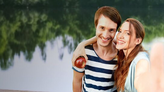Lächelndes Paar posiert am Ufer eines Sees und hält gemeinsam einen Apfel in der Hand.