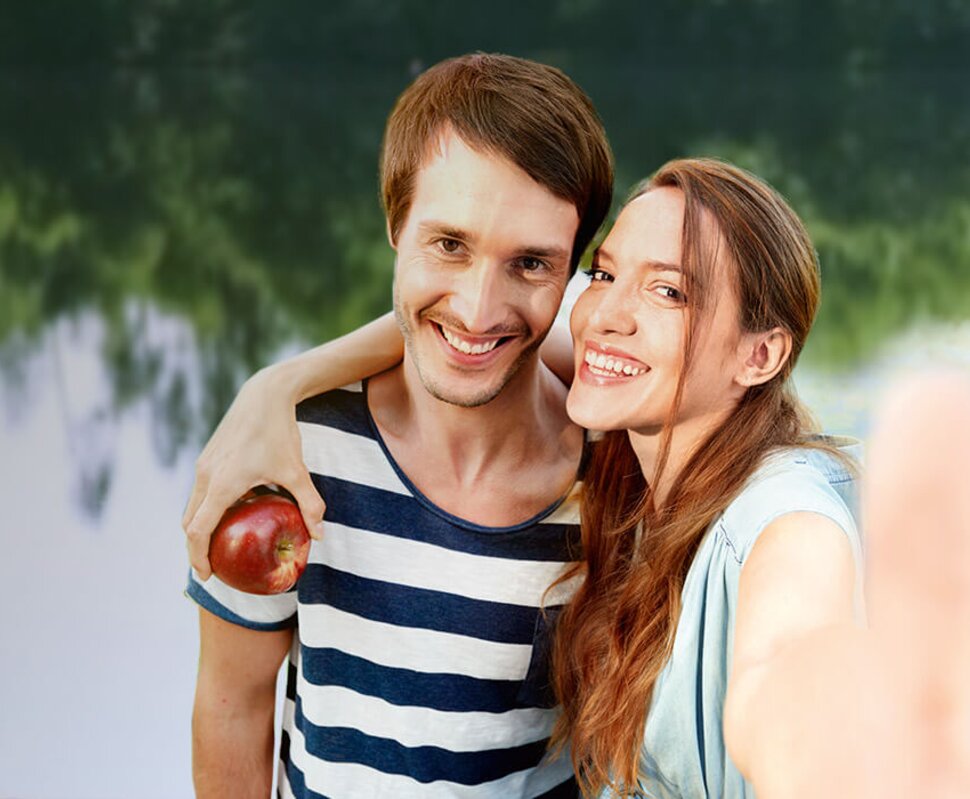 Lächelndes Paar posiert am Ufer eines Sees und hält gemeinsam einen Apfel in der Hand.