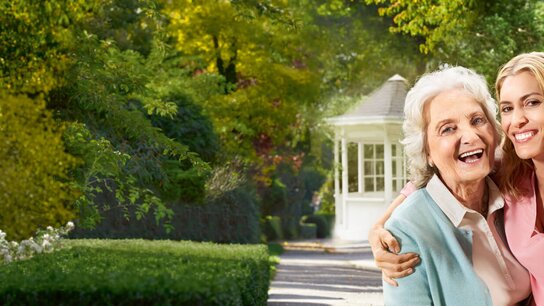 Zwei lächelnde Frauen posieren im Garten für ein Selfie, umgeben von grünen Bäumen und einem weißen Pavillon.