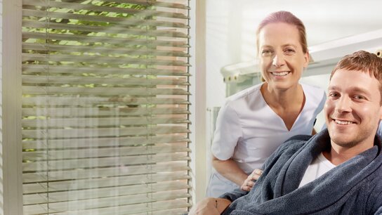 Ein lächelnder Patient in einem Bademantel sitzt neben einer Pflegekraft in einem hellen Raum mit Jalousien.