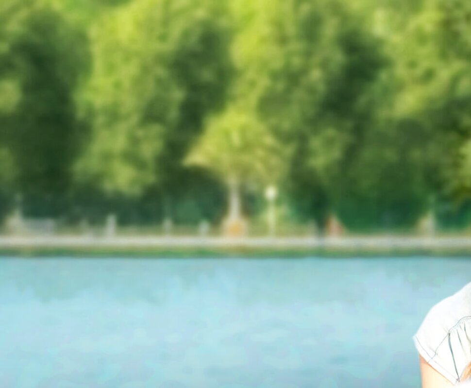 Lächelnde Frau im Vordergrund, steht am Wasser und blickt freundlich zu einer Person außerhalb des Bildes. Grünes Ufer im Hintergrund.