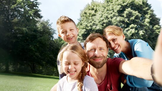 Familie posiert lachend für ein Selfie im Freien, umgeben von Bäumen und Grün. Kinder und Eltern strahlen Freude aus.