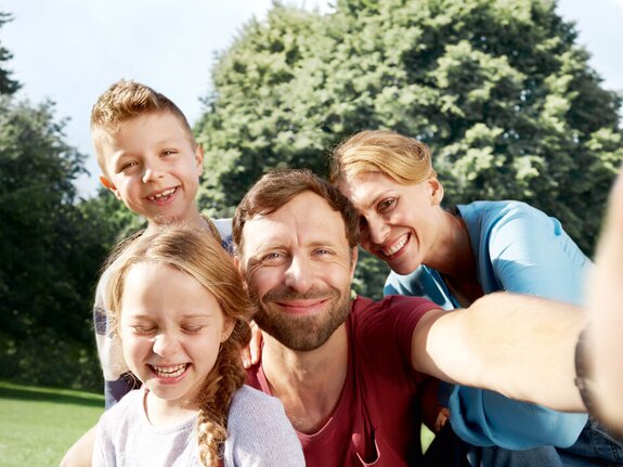 Familie posiert lachend für ein Selfie im Freien, umgeben von Bäumen und Grün. Kinder und Eltern strahlen Freude aus.